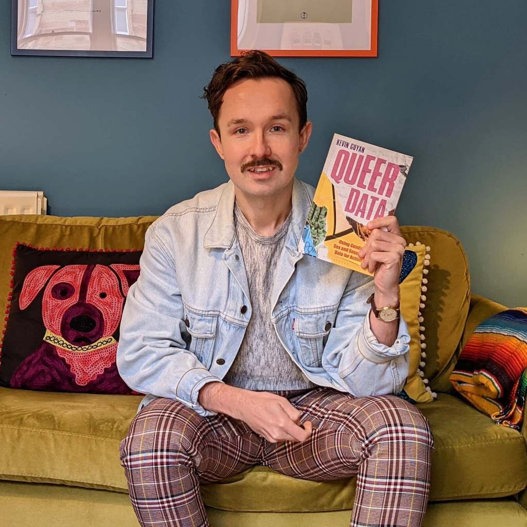 Kevin is sitting on a green sofa, in his left hand is a copy of the book Queer Data. He is wearing a denim jacket.