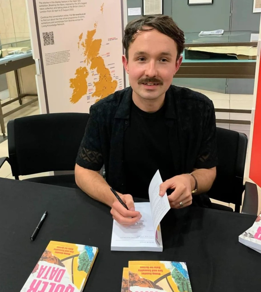 Kevin is sitting at a signing desk at a book event. He is wearing a short-sleeved black top and signing copies of Queer Data.