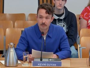 Kevin wearing a blue jacket in front of a microphone and sign with his name. He is speaking at a committee session at the Scottish Parliament.