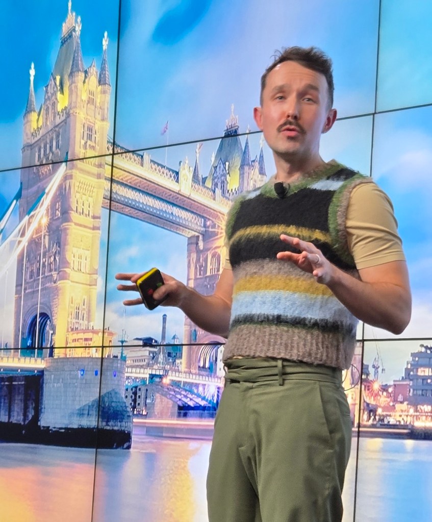 Kevin is delivering a talk. He is wearing a light brown t-shirt with a wooly vest. He is also wearing olive green trousers. He is standing in front of a screen with London's Tower Bridge on display.
