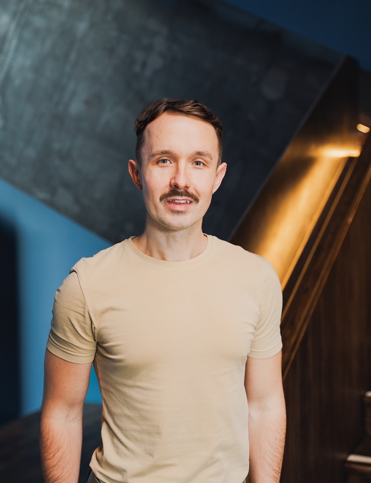 Kevin Guyan, a white man with dark brown hair and a moustache. He is wearing a light brown t-shirt and standing in front on a backdrop with industrial colours of brown, blue and grey.