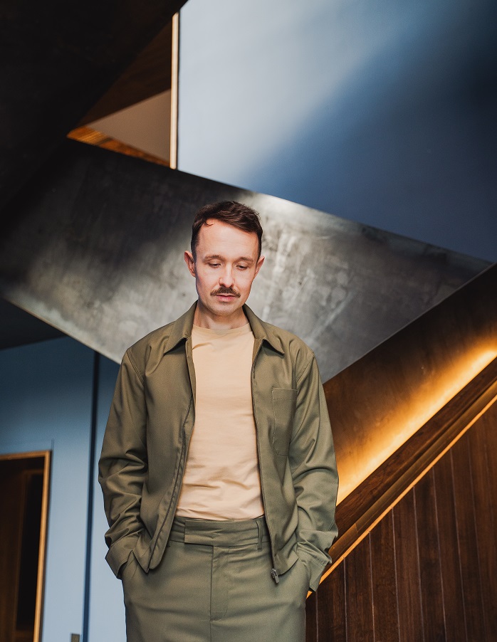 Kevin is standing in front of an industrial, metallic staircase. He is wearing a light brown t-shirt and a green blazer jacket.